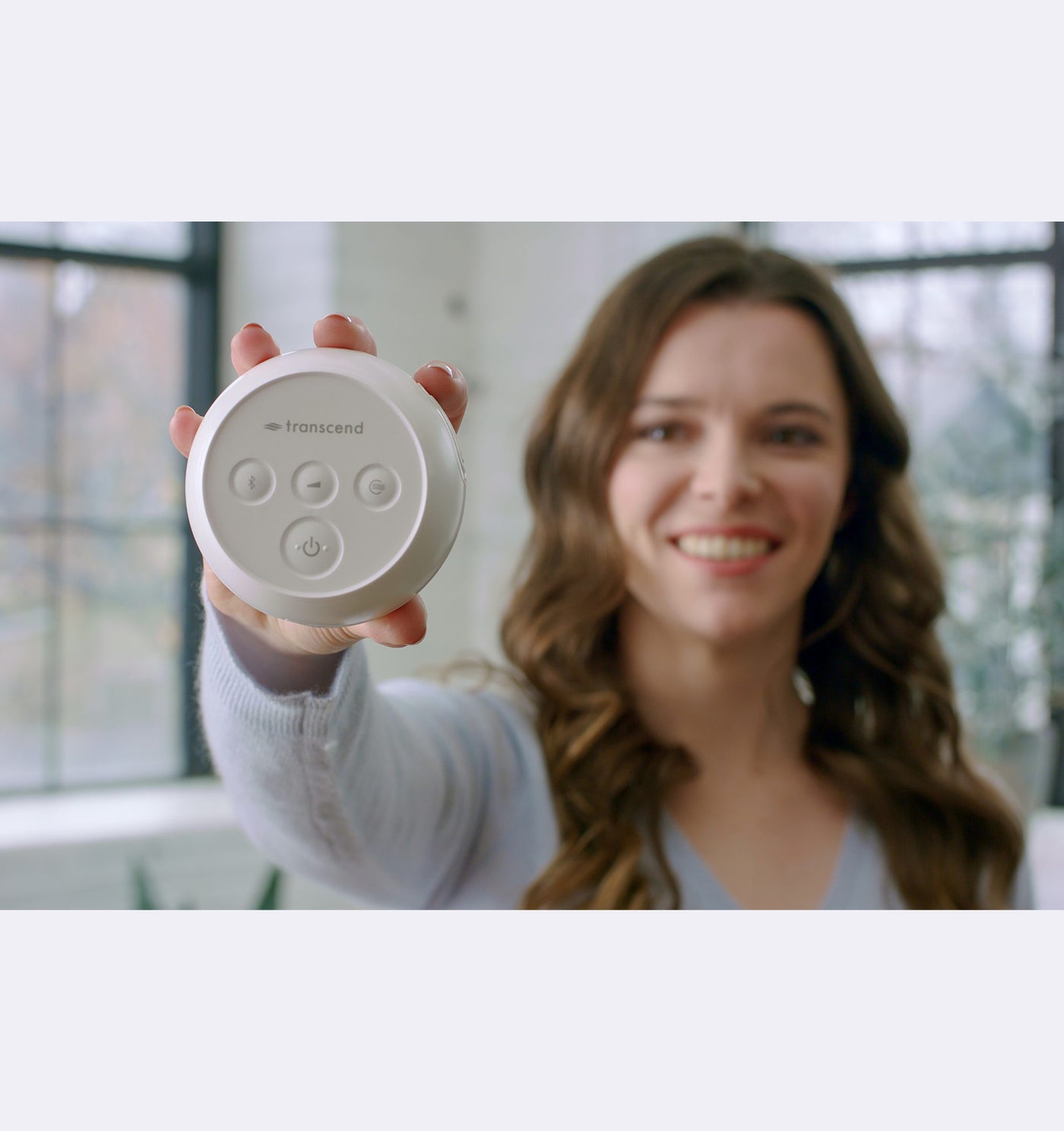 Woman holding a white device with buttons, smiling in a room with large windows.