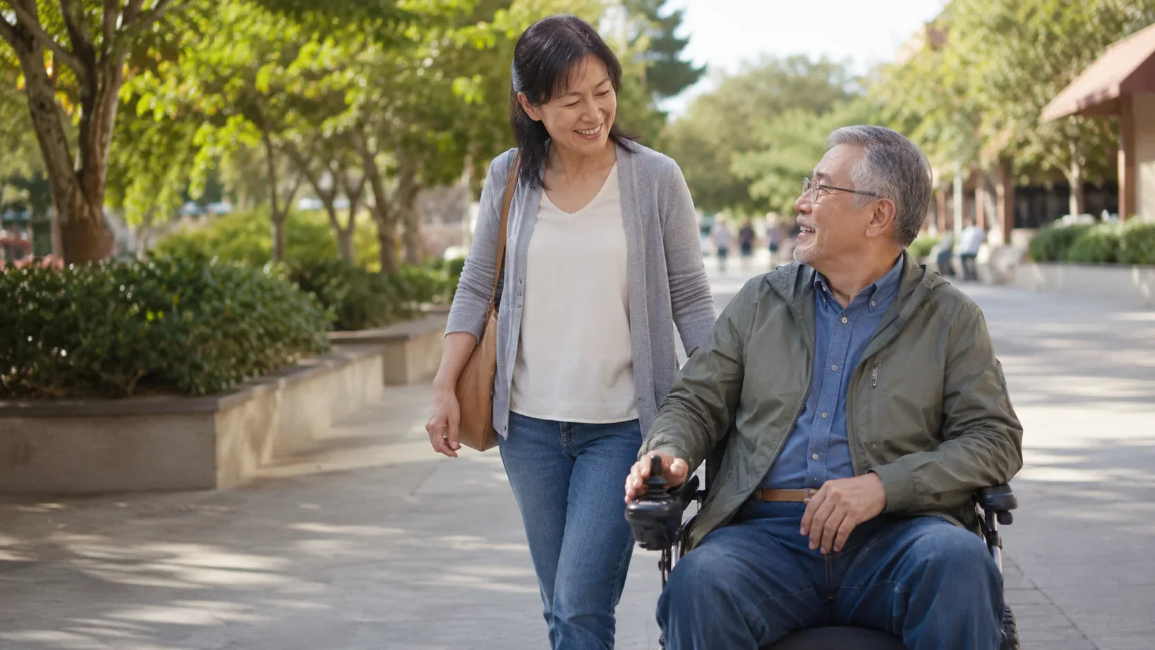 Older man using a power wheelchair while walking outdoors with a loved one in a peaceful park setting.