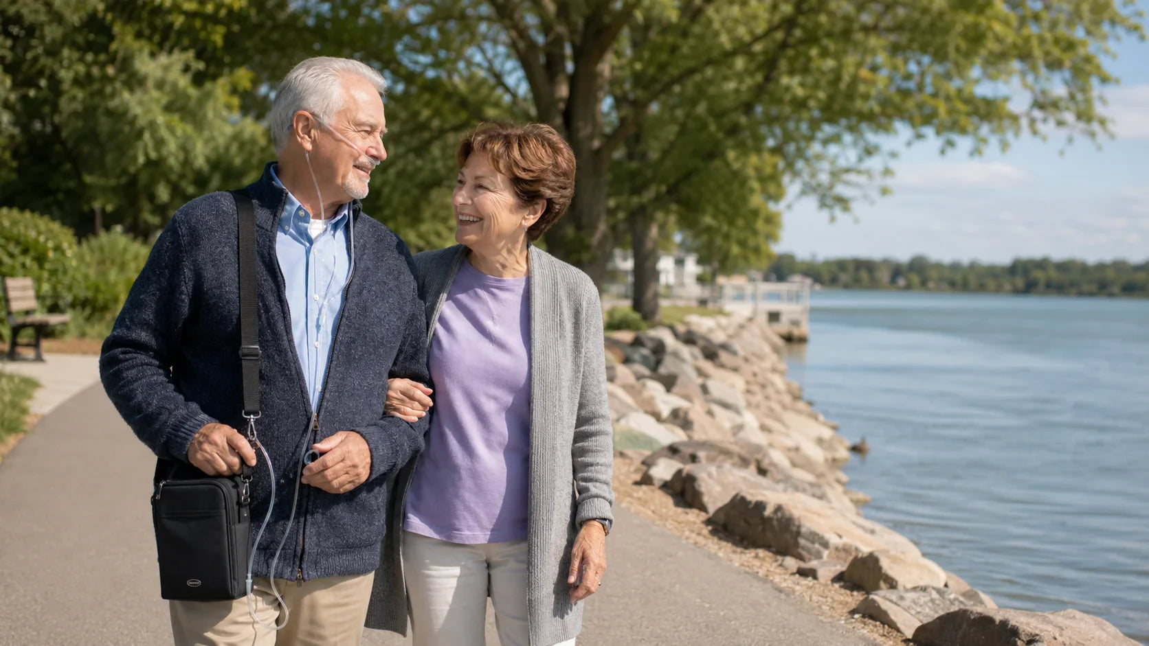 Older man wearing a nasal cannula and carrying a portable oxygen concentrator while walking by the water with his wife.