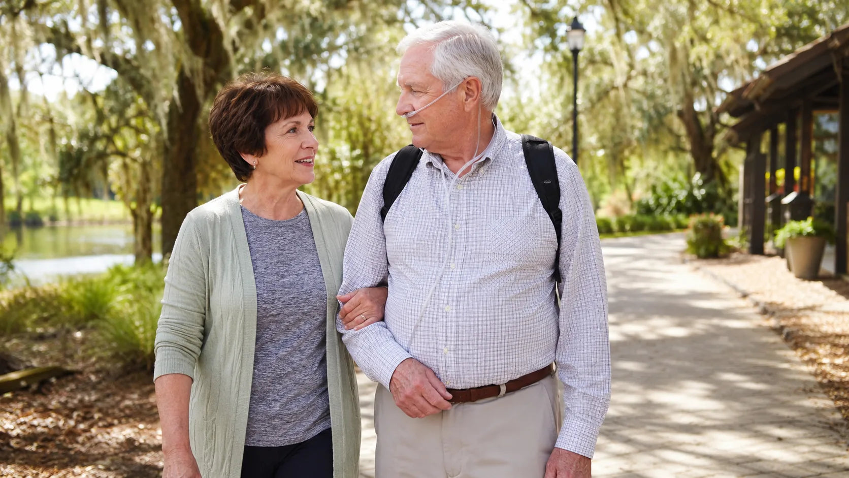 Older man wearing a nasal cannula and carrying a portable oxygen concentrator while walking outdoors with his wife.
