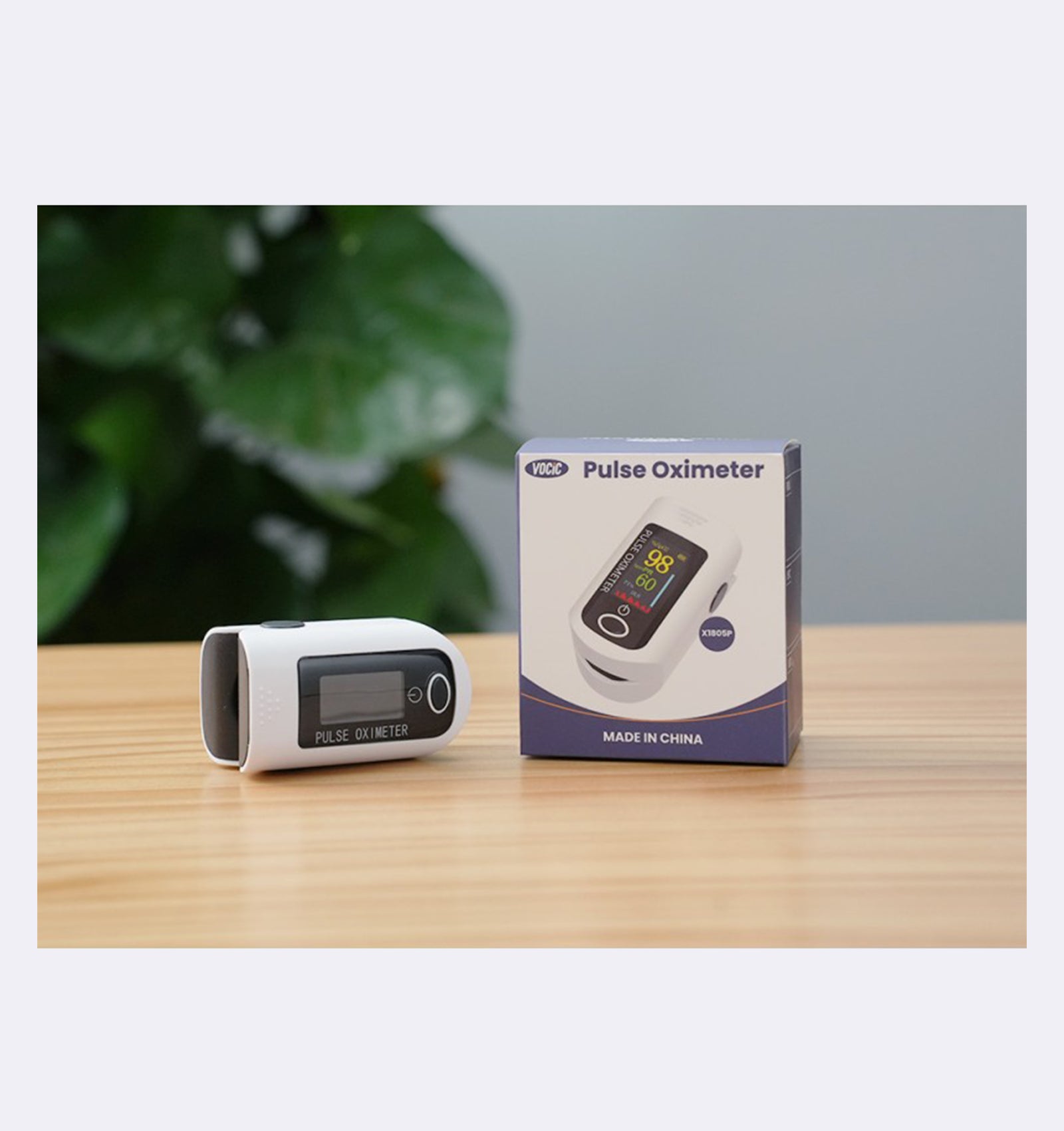 Pulse oximeter and its packaging on a wooden surface with a blurred green plant in the background