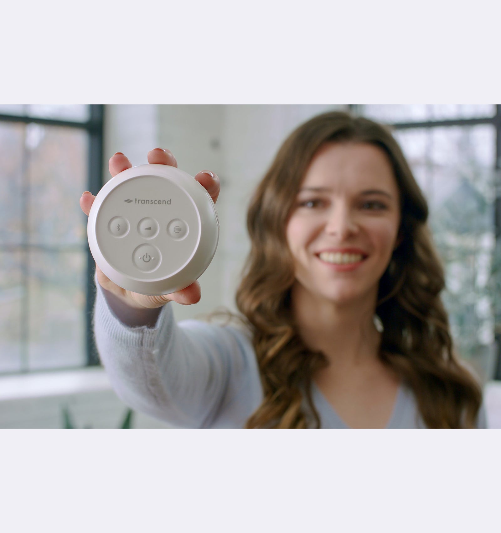 Woman holding a white device with buttons, smiling in a room with large windows.